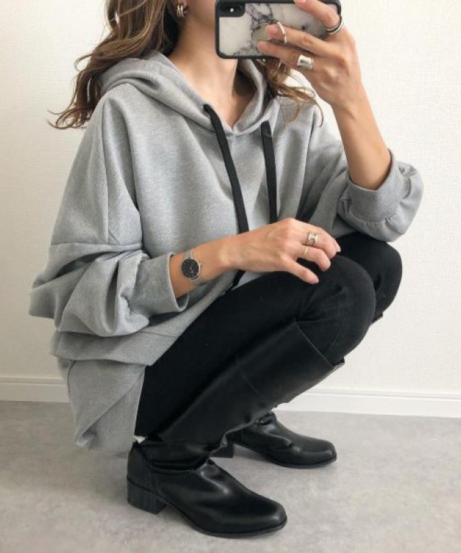 グレーのオーバーサイズフーディーを着用した女性が、黒のスキニーパンツとロングブーツを合わせ、座っている全身ショッ