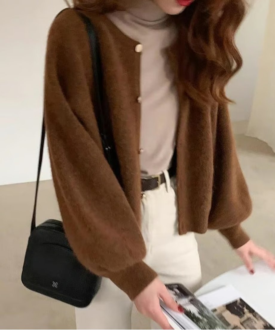 コーヒーのランタンスリーブショートカーディガンと淡いベージュのタートルネック、白いパンツを着用した女性のバストアップ。黒いショルダーバッグを肩掛け。