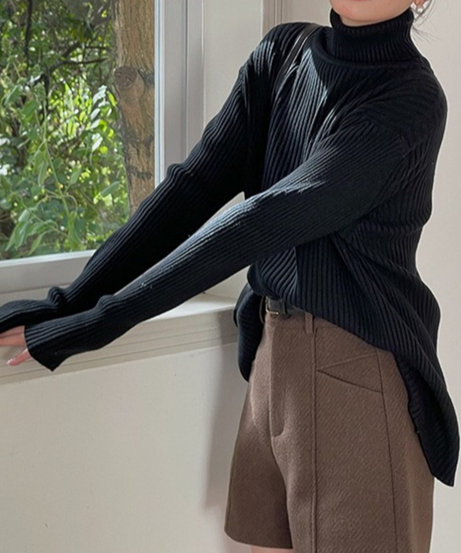 ブラックのリブ編みタートルネックセーターを着用し、茶色のショートパンツを合わせた女性の上半身。窓際で撮影され、セーターの裾の前後差が確認できる。