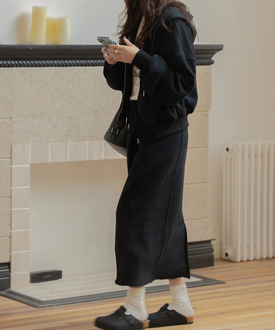黒のスウェットジップアップパーカーとロングスカートをセットアップで着た女性の横からのスタイル。手にスマホを持っている。