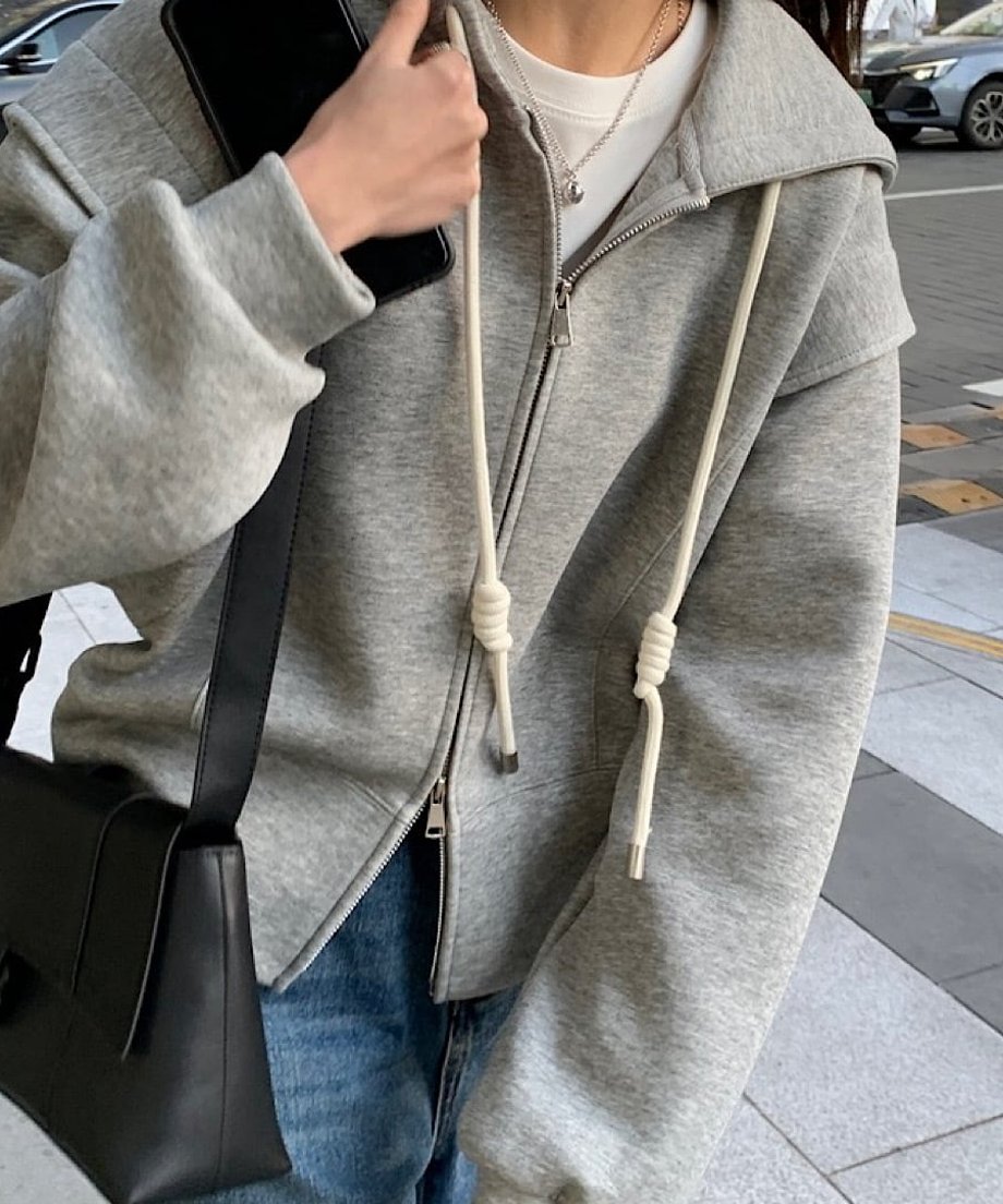 グレーのダブルジップデザインフーディを着用し、バッグを肩にかけた女性の胸元付近のクローズアップ。白のインナーとネックレスを合わせた様子。