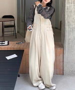 屋外ので立ちポーズをとる女性。ベージュのバギーワイドオーバーオールに、黒と白のボーダーのシアー長袖Tシャツをインナーに着用。白いスニーカーとベージュのトートバッグを合わせている。
