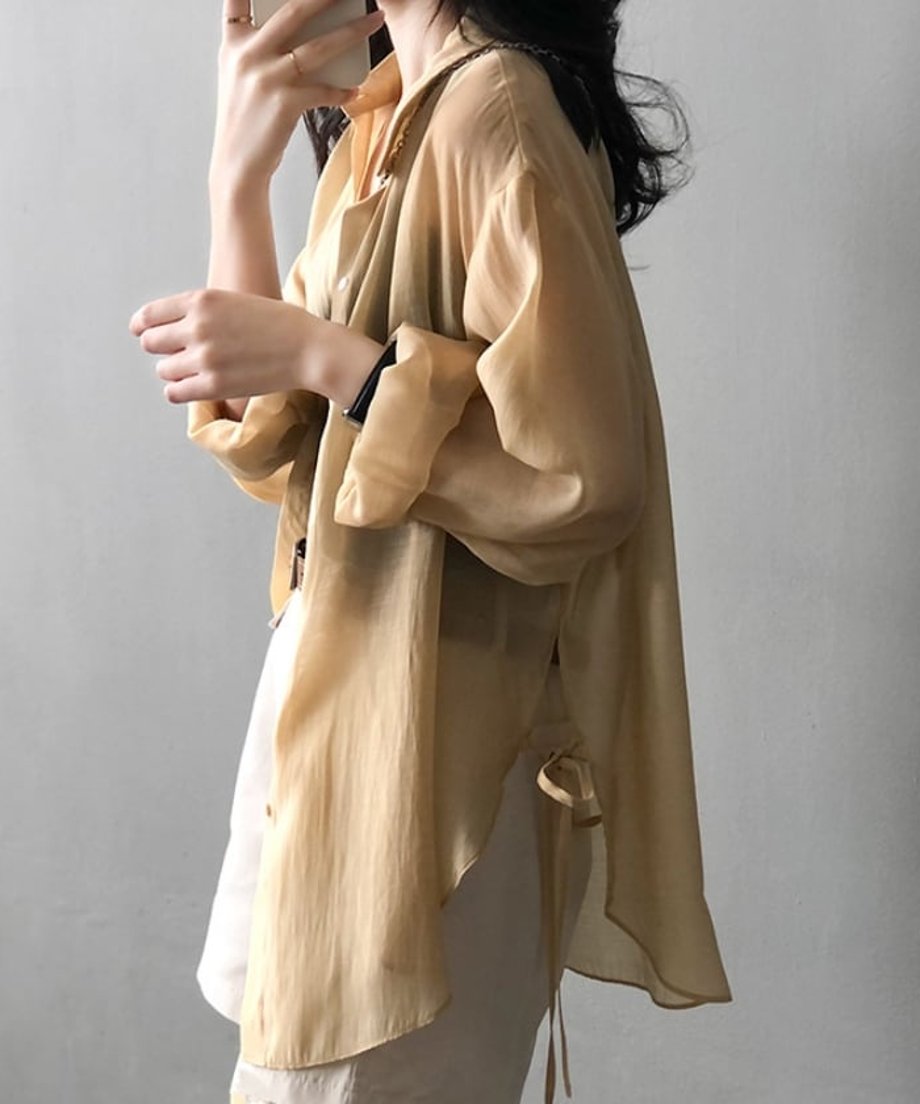 イエローのシアーシャツを着用し、袖を捲り上げて鏡越しに自撮りをする女性