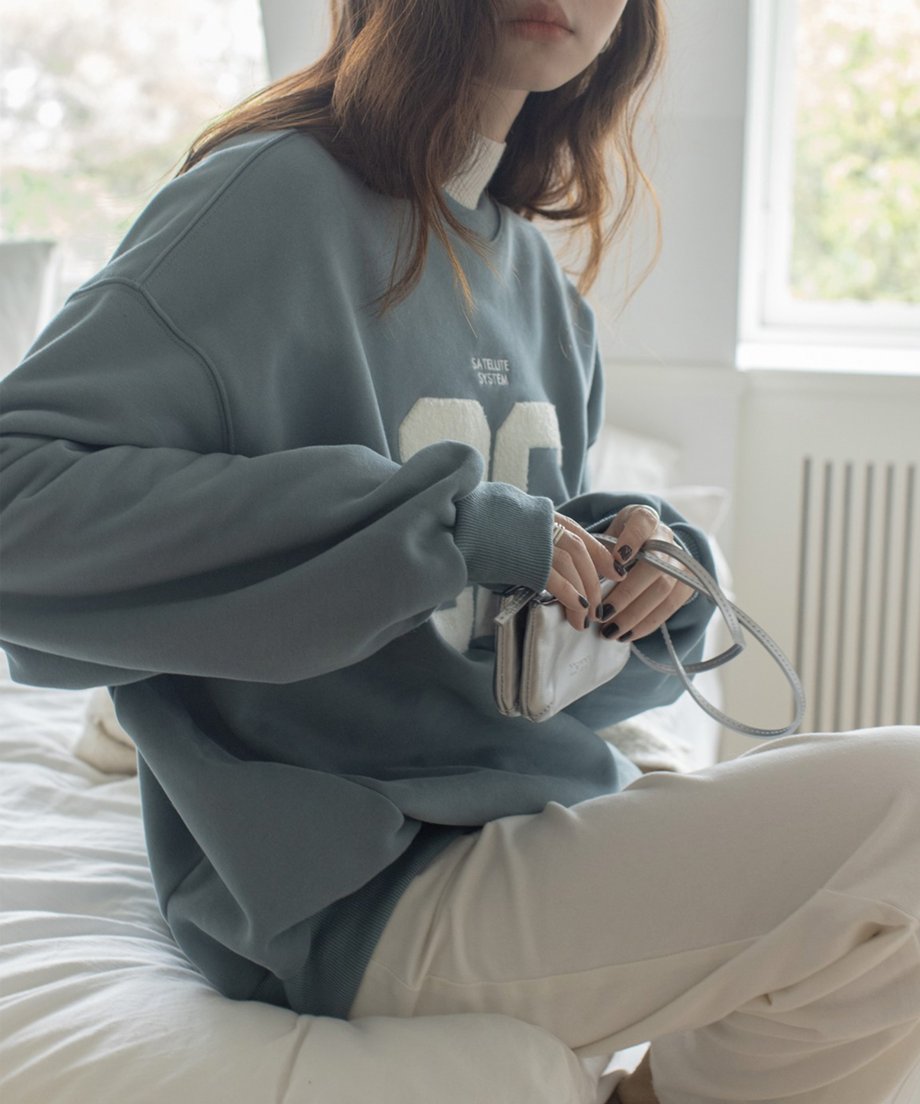 ミストブルーのスウェットシャツを着た女性がベッドに座りバッグを持っている様子。