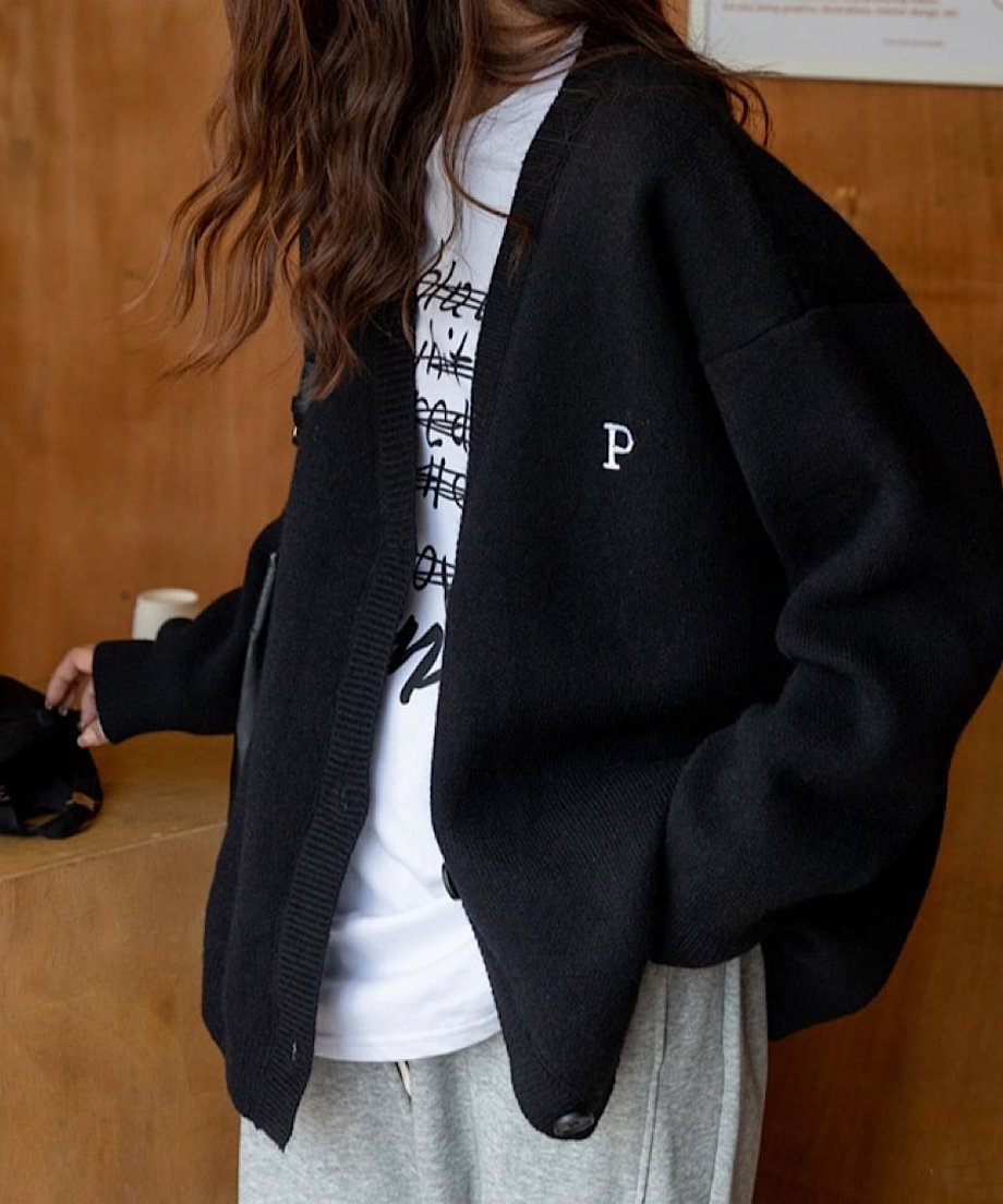 黒いオーバーサイズカーディガンの左胸にある白い「P」の刺繍ロゴとボタンをクローズアップした写真。