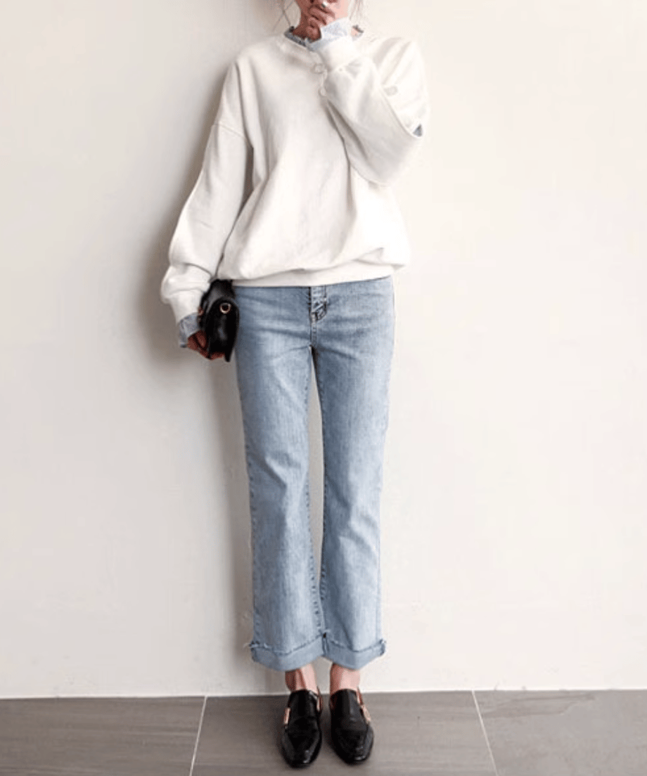 ホワイトのレイヤードスウェットを着用した女性の全身画像。フリルネックと袖口からブルーストライプの生地が覗く重ね着風デザイン。薄いブルーのフレアデニムと黒のローファーを着用。