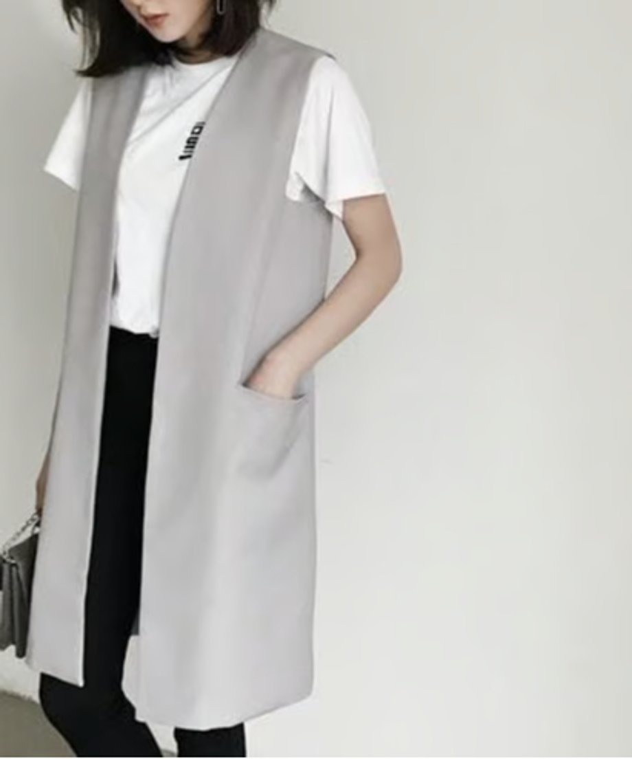 白いTシャツと黒いパンツにライトグレーのロングベストを着用した女性が、片手をポケットに入れ、バッグを持っている。