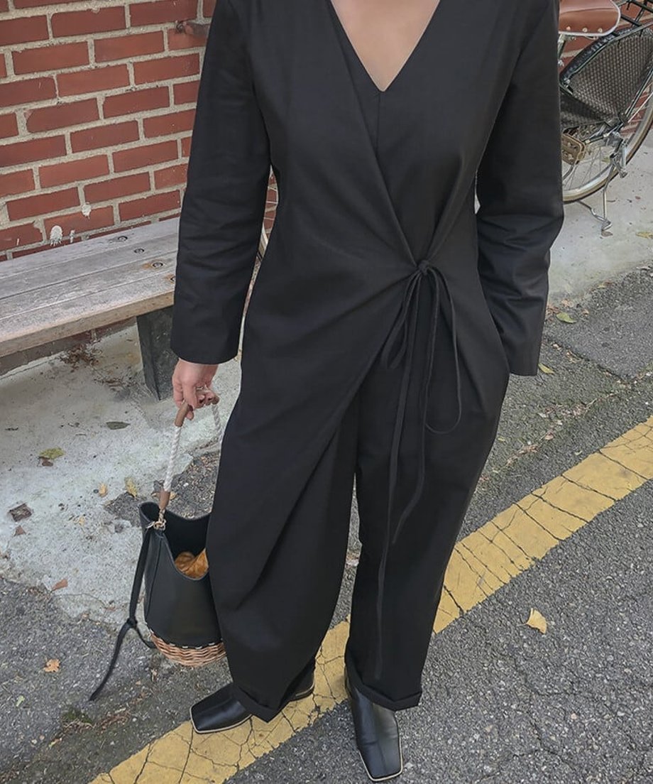 黒のVネックラップオールインワンを着た女性のウエストから上のクローズアップ。ウエストの結び目がはっきりと見え、左手に黒と籐のバッグを持っている。