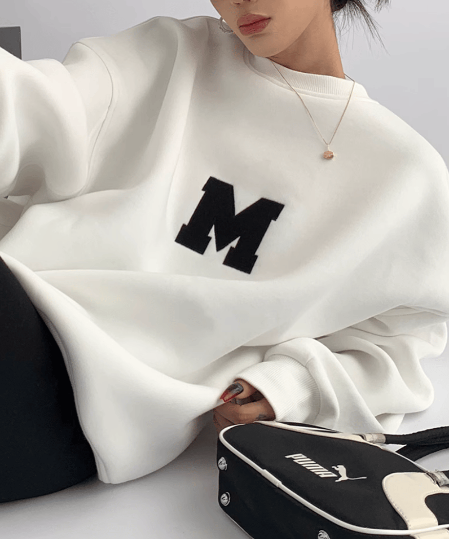 「M」が施されている白いオーバーサイズスウェットを着用した女性が横になっている。黒のバックが横に置かれたショット。