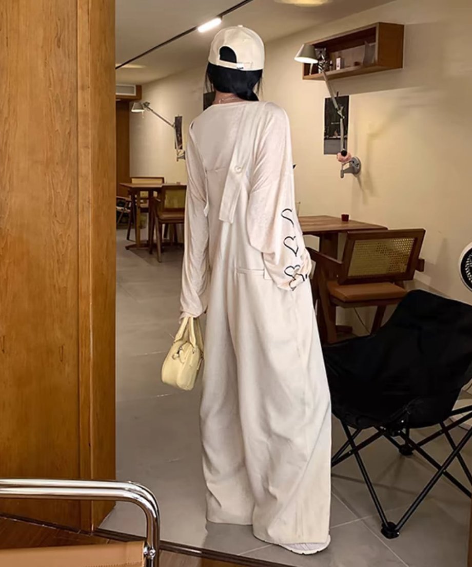 カフェのような場所で後ろを向いて立つ女性の全身写真。ベージュのオーバーオールと長袖Tシャツの組み合わせ。Tシャツの背中側のロゴと、キャップ、ミニバッグが確認できる。