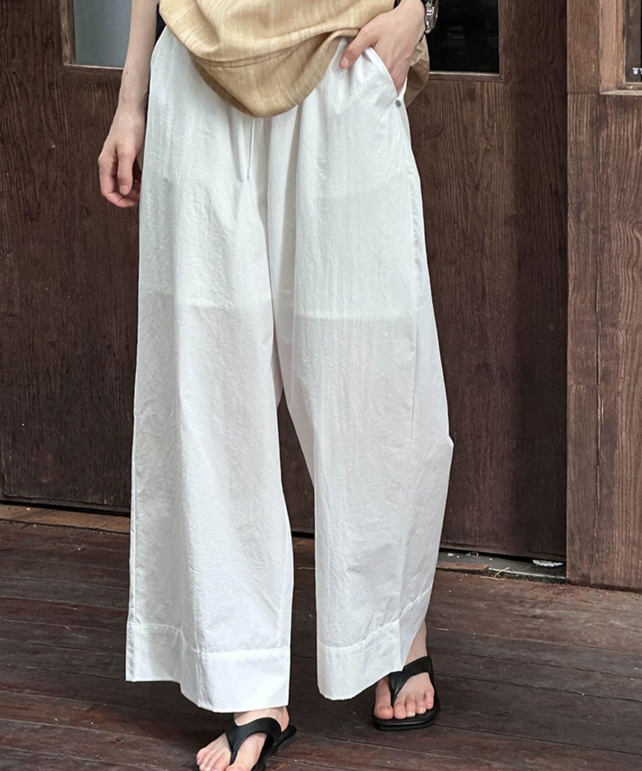 ホワイトのワイドレッグパンツとベージュのトップスを着用し、黒のビーチサンダルを履いた女性の下半身正面。ポケットに手を入れている。