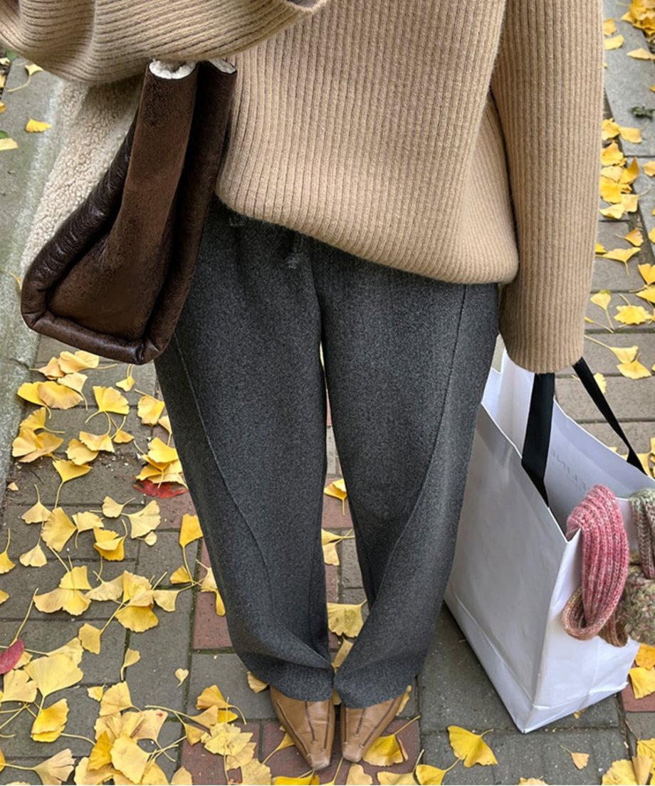 ダークグレーのセンターシームワイドパンツを着用した女性が、イチョウの落ち葉の上に立っているクローズアップ。キャメル色のニットとムートン素材のバッグを合わせている。