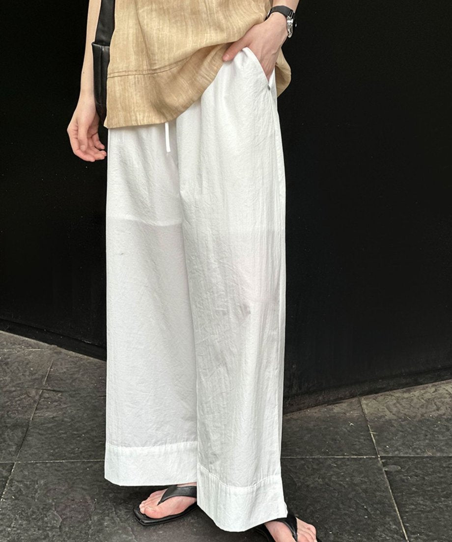ホワイトのワイドレッグパンツとベージュのトップスを着用し、黒のバッグを持った女性の下半身側面。黒のビーチサンダルを履いている。