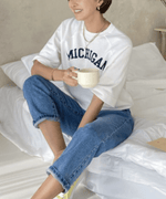 ホワイトのロゴTを着た女性がベッドに座り、リラックスしてコーヒーを飲んでいる。