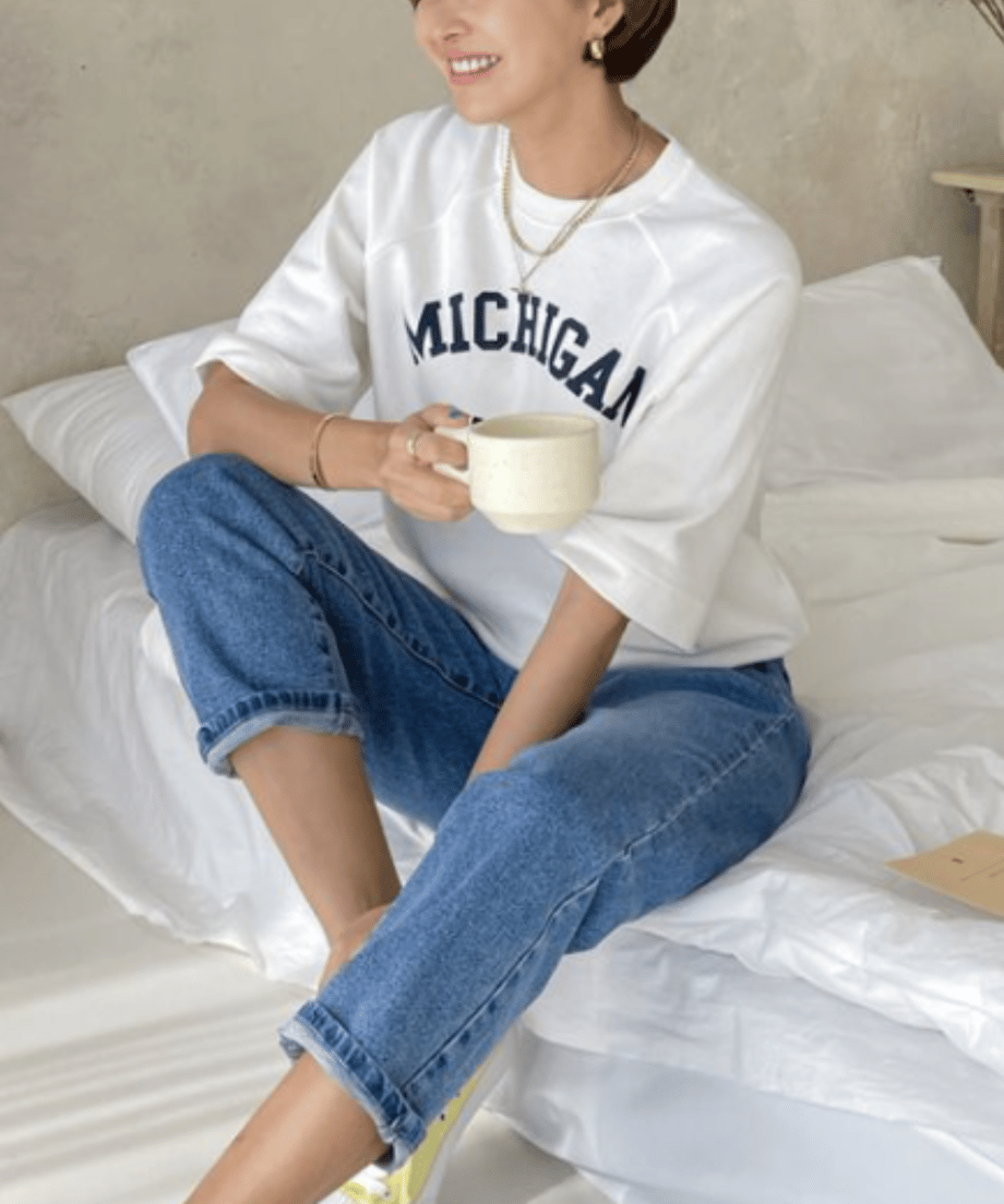 ホワイトのロゴTを着た女性がベッドに座り、リラックスしてコーヒーを飲んでいる。