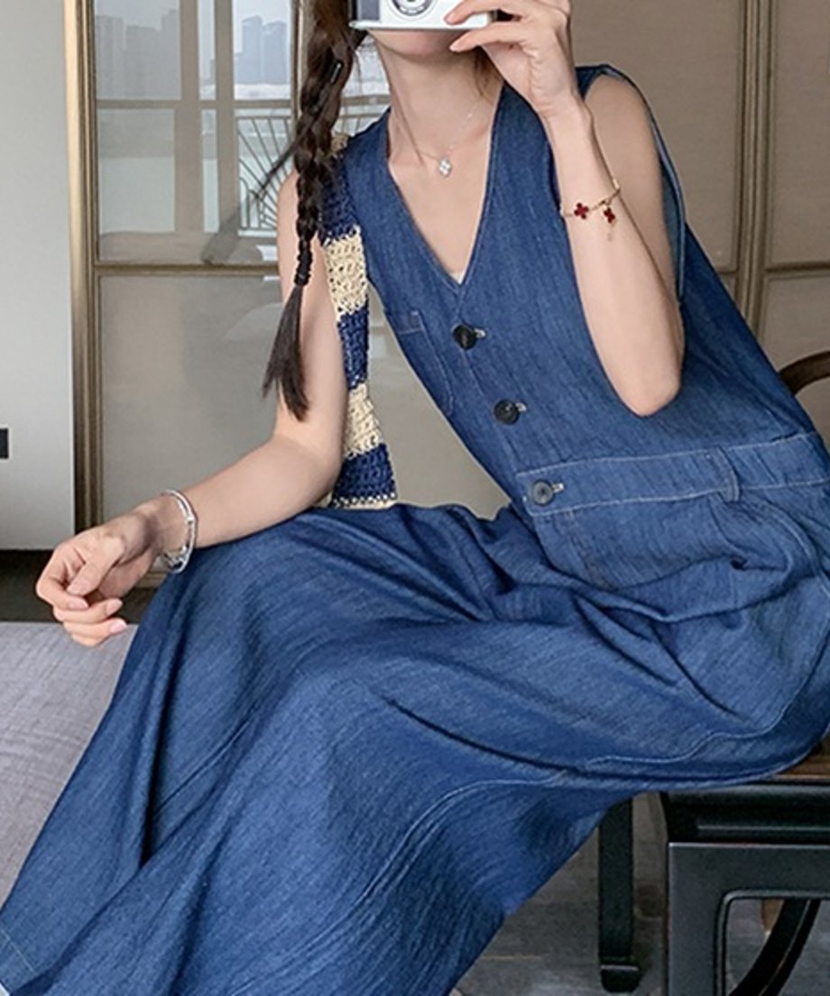 Vネックのデニムワイドレッグジャンプスーツを着用し、座っている女性のバストアップ写真。太めの三つ編みと編みバッグ、華奢なネックレスがポイント。