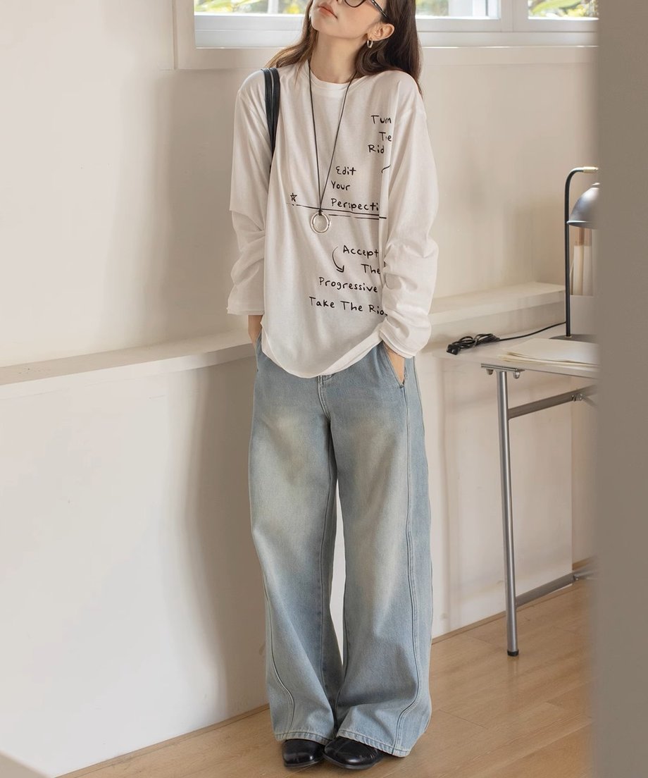 白のメッセージプリントロングTシャツとライトブルーのワイドデニムパンツを着用した女性の全身。ゆったりとしたシルエットで、黒のショルダーバッグとローファーを合わせたカジュアルスタイル。