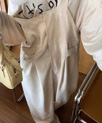 カフェのような場所での女性のウエストアップ。ベージュのオーバーオールとロゴ入りTシャツの組み合わせ。オーバーオールのウエストのタックとサイドのベルトディテールが確認できる。