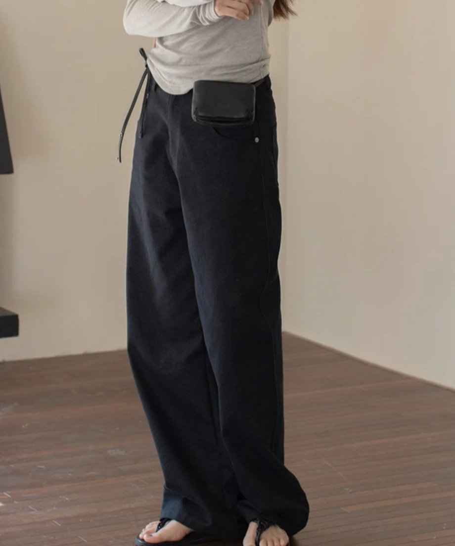グレーのトップスとブラックのワイドレッグパンツを着たモデルが部屋で立っている画像。シンプルでスタイリッシュなコーディネートが可能。