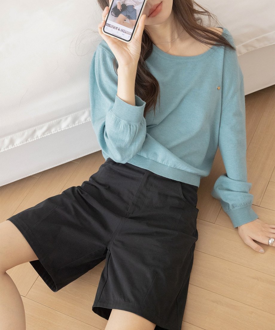 グラスブルーのUネックニットと黒のショートパンツを着用した女性が、床に座りスマートフォンを見ている様子。袖口に少しボリュームのある、リラックスした夏のカジュアルスタイル。