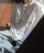 白のメッセージプリントロングTシャツとワイドパンツを着用し、窓辺に座っている女性。デザイン性のあるネックレスをしている。