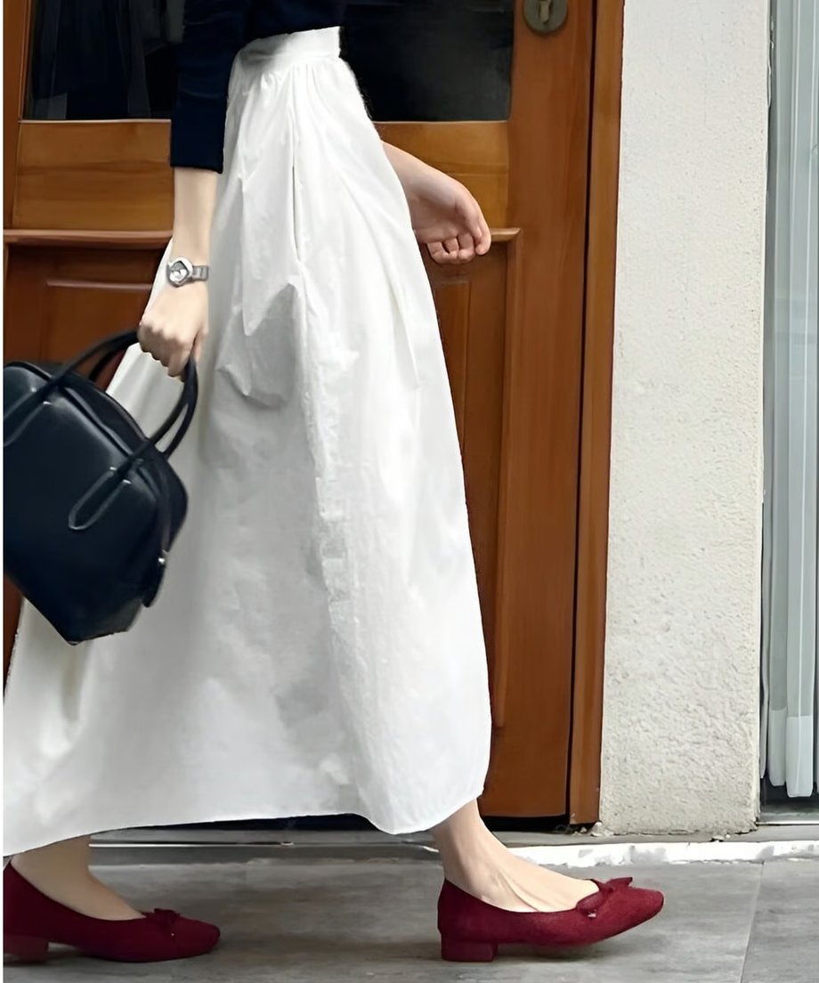 白いマキシ丈のギャザースカートにボルドーのフラットシューズを合わせた女性の歩行シーン。