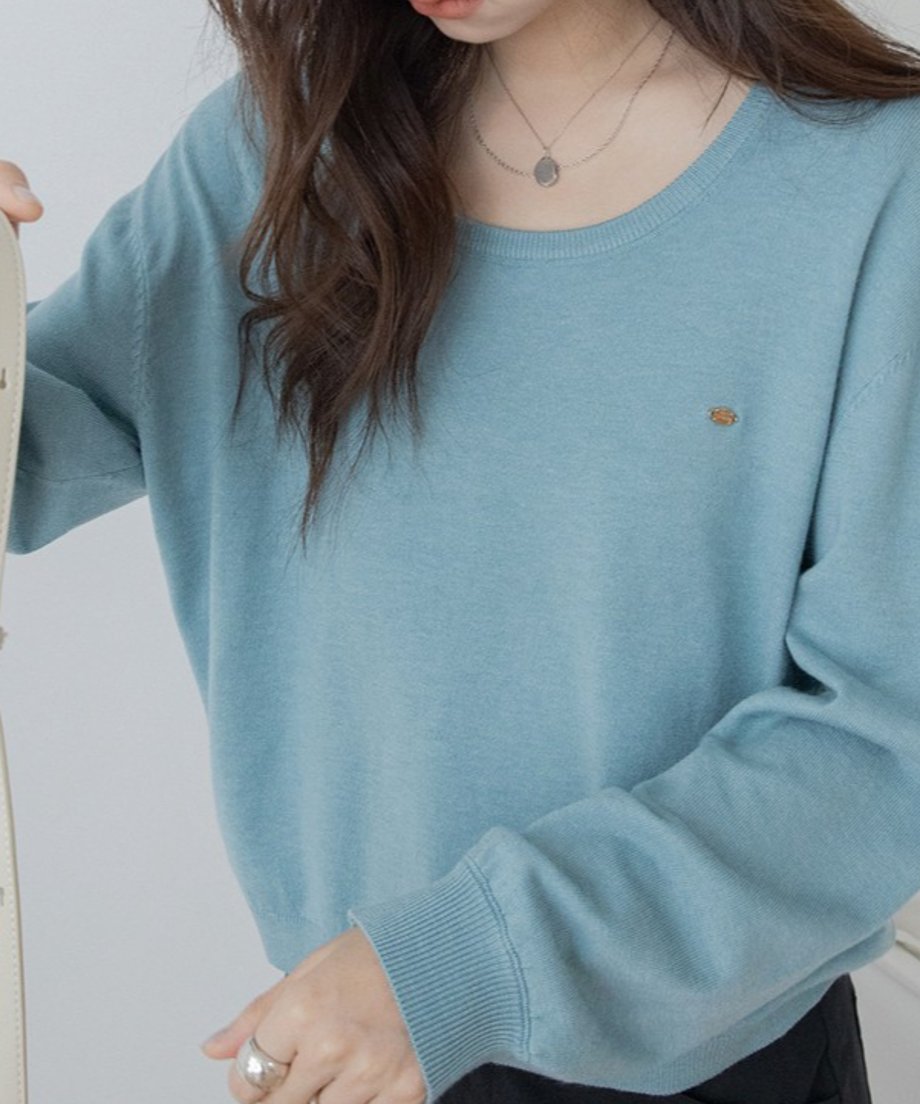 グラスブルーのUネックニットを着用した女性のバストアップ。控えめなワンポイントワッペン、リブ編みの袖口が確認できる、春らしいきれいめな着こなし。