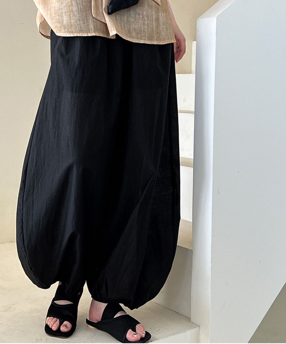 ブラックのバルーンパンツ（裾絞り）とベージュのトップスを着用し、屋内の階段に立つ女性の、下半身側面