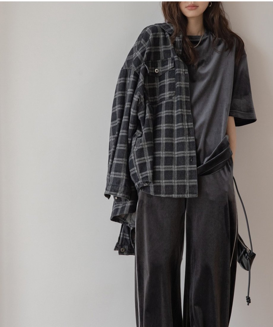 ダークグレーのベルベット素材のセットアップを着用した女性。黒のチェック柄のシャツを肩にかけ、ミニバッグを提げている。トップスはオーバーサイズTシャツ、ボトムスはサイドにパイピングが入ったワイドパンツ。