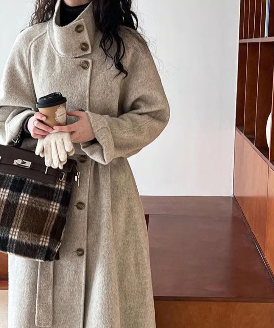 モカのスタンドカラーコートを着用した女性のバストアップ。茶色のチェック柄バッグとコーヒーカップを持っている。