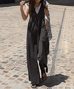 グレーのワイドレッグサロペットを着た女性が、白Tシャツをインナーに着用し、柄物の大きなバッグを肩にかけ、サンダルを履いて石畳の上を歩いている全身ショット。