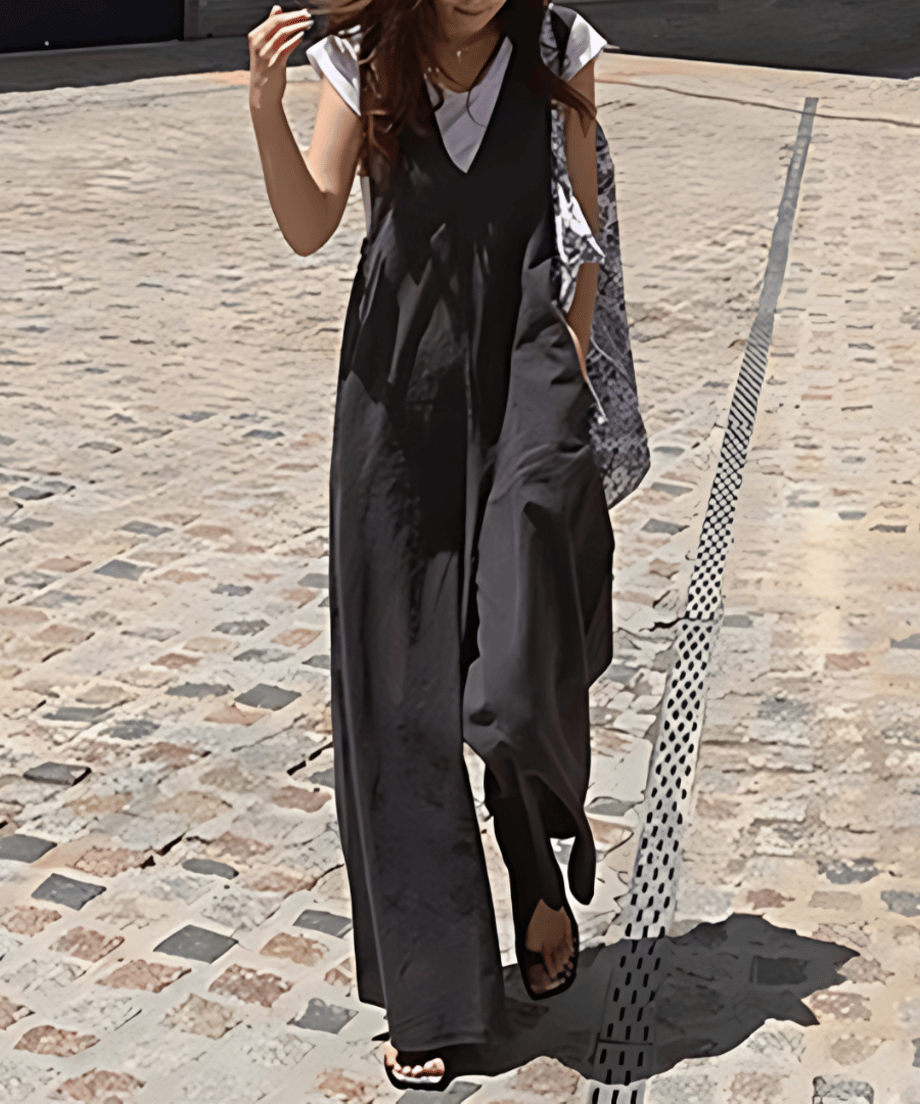 グレーのワイドレッグサロペットを着た女性が、白Tシャツをインナーに着用し、柄物の大きなバッグを肩にかけ、サンダルを履いて石畳の上を歩いている全身ショット。