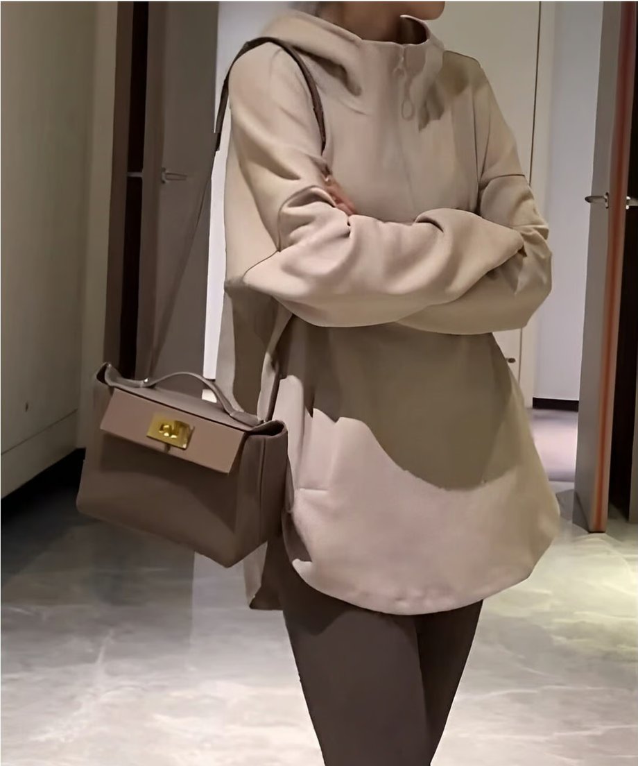 オフホワイトのオーバーサイズフーディーを着用し、茶色のレギンスを合わせた女性が、斜めがけのミニバッグを持って立っている。