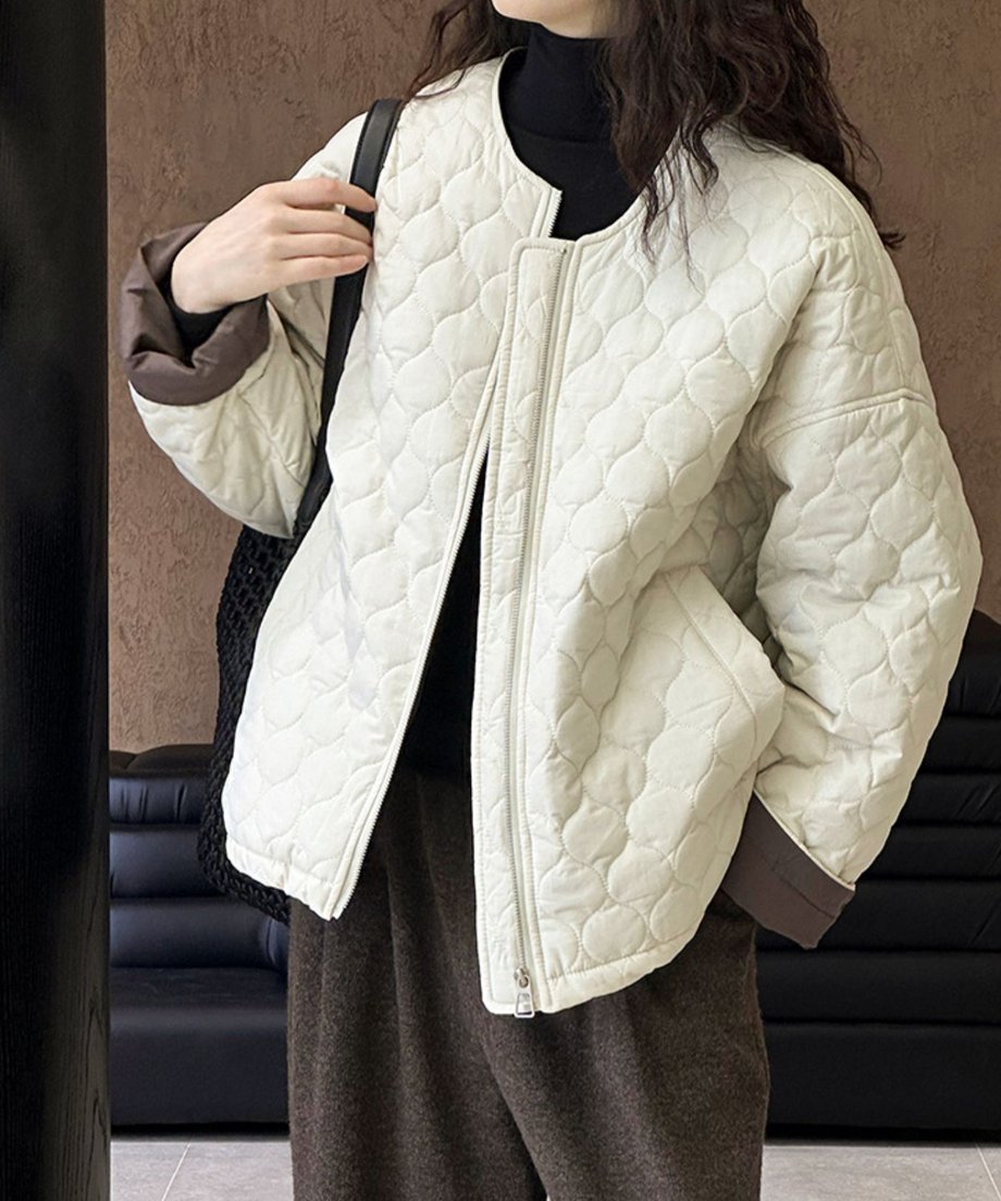 ホワイトのカラーレスのキルティングジャケットを前開きで着用した女性。黒のタートルネックとダークブラウンのボトムス、黒いバッグを合わせた全身像。