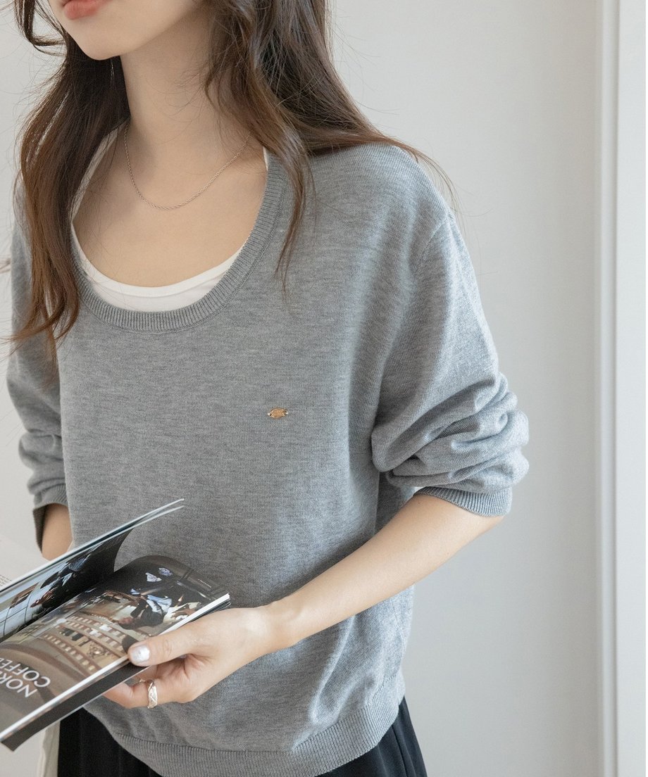 グレーのUネックニットを着用した女性のバストアップ。小さな惑星の刺繍と、インナーの白Tシャツ、手元に雑誌を持つ様子。