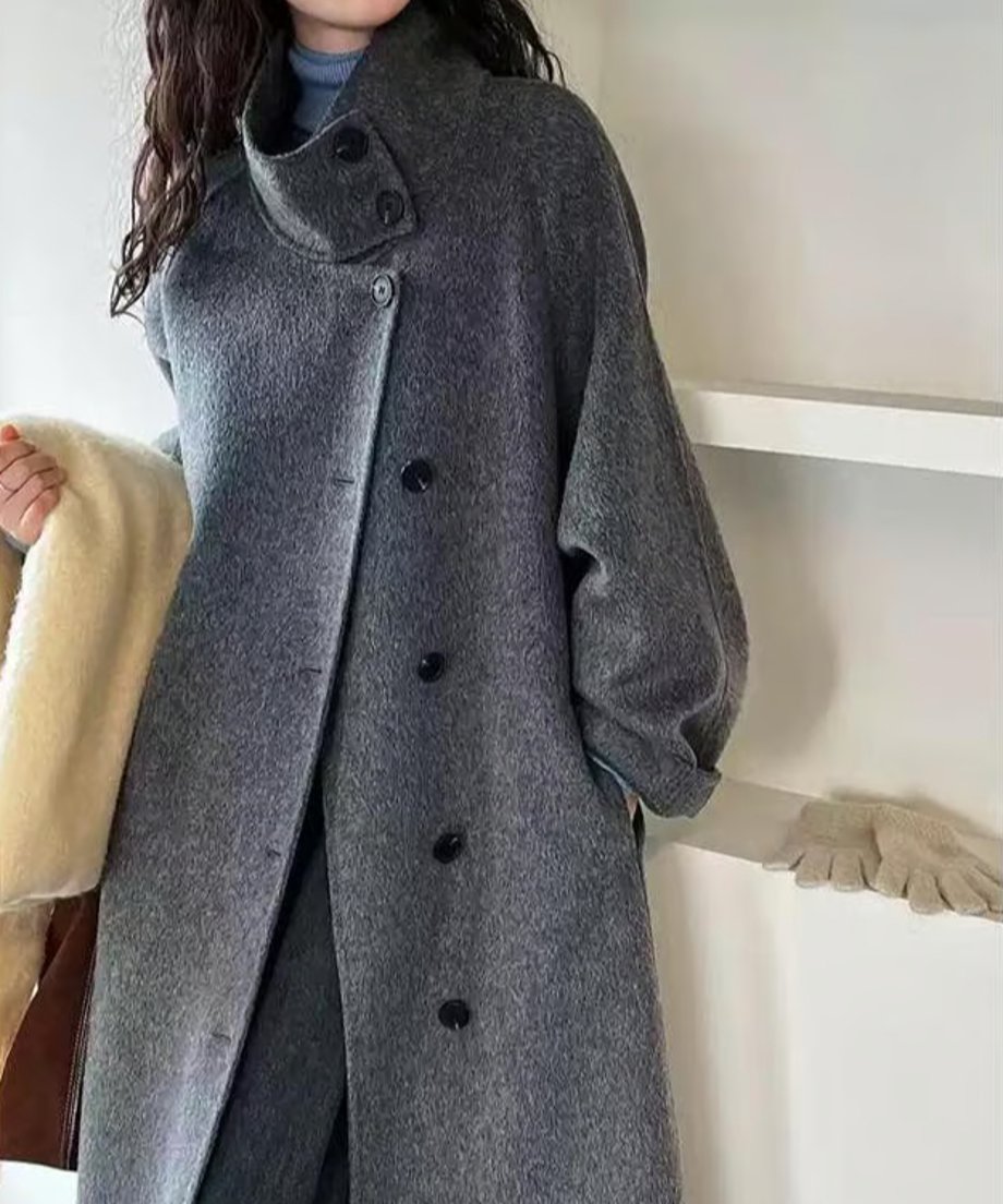 ダークグレーのスタンドカラーコートを着用した女性の上半身。襟元のボタンと、アイボリー色のマフラーを手に持っている。
