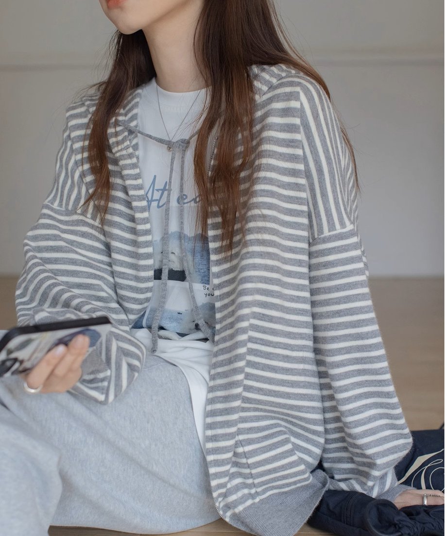 グレーと白の細いボーダー柄のジップアップパーカーを着用した女性が座っている半身。インナーには白いTシャツ、ボトムスにはライトグレーのスウェットパンツを合わせたカジュアルな着こなし。