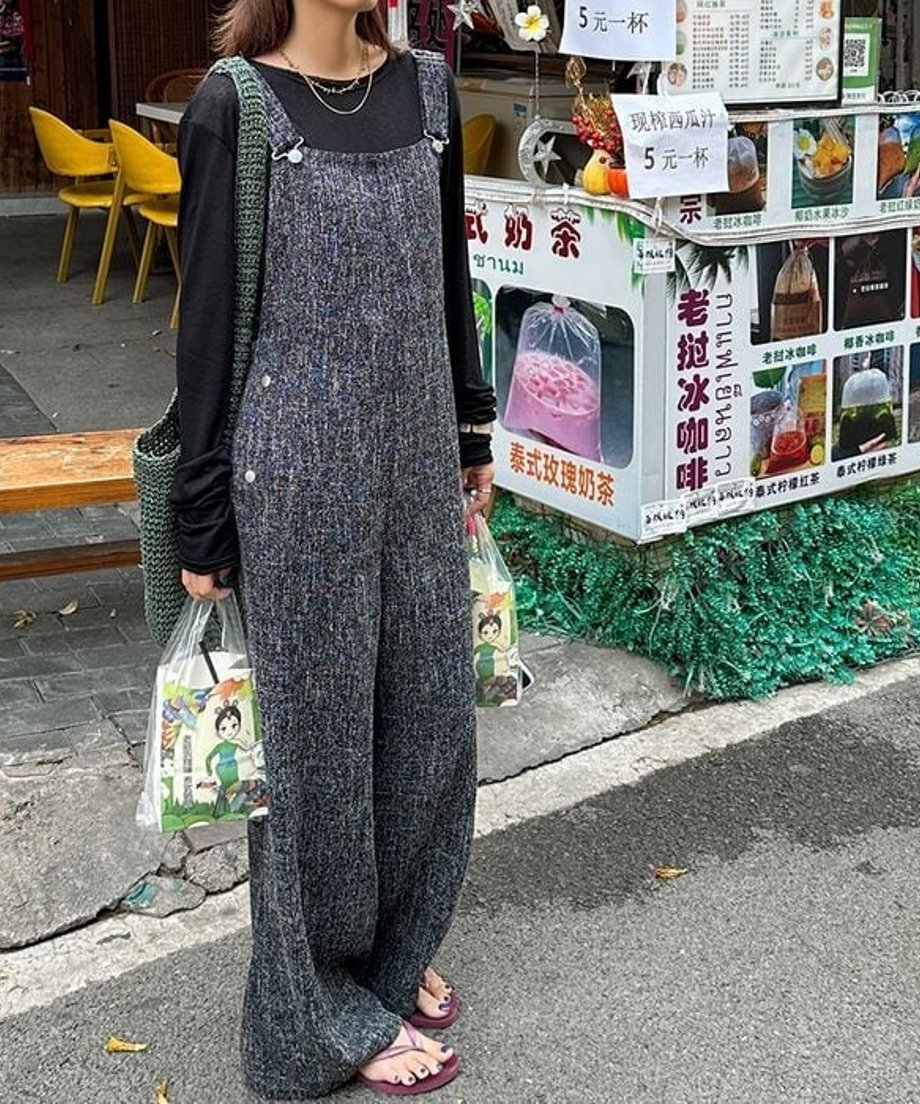 ブラックのワイドレッグオーバーオールを着用し、街角の売店前に立つ女性の全身カット。