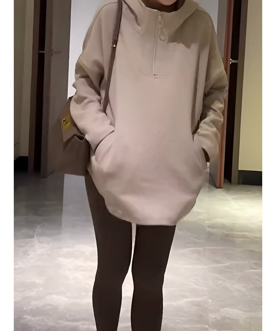 オフホワイトのハーフジップフーディーを着用し、両手をポケットに入れ正面を向いている女性。裾は丸くカットされており、茶色のレギンスと合わせている。