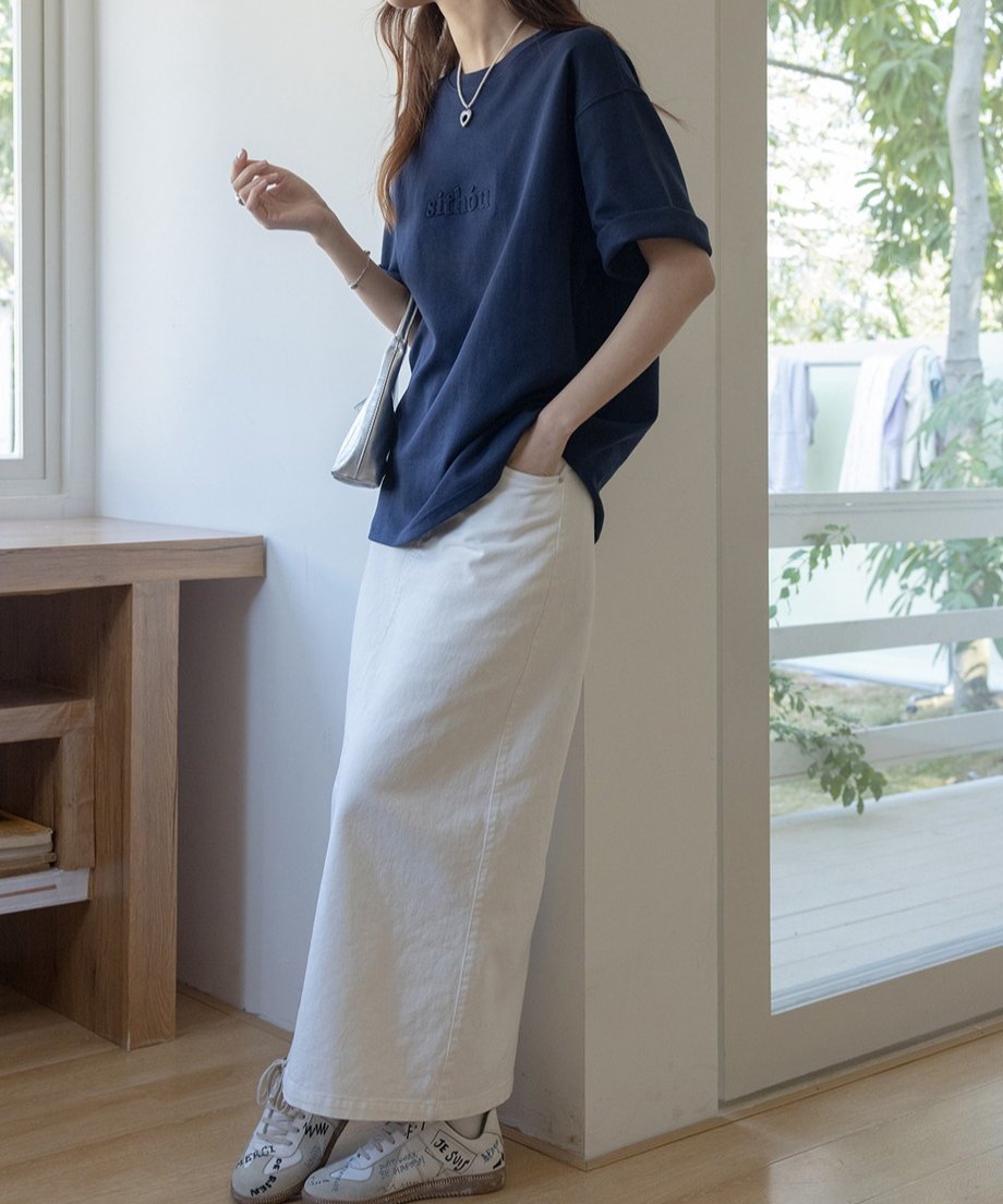 ネイビーのロゴTシャツを着た女性が、室内でリラックスした様子。白いスカートとコーディネート。
