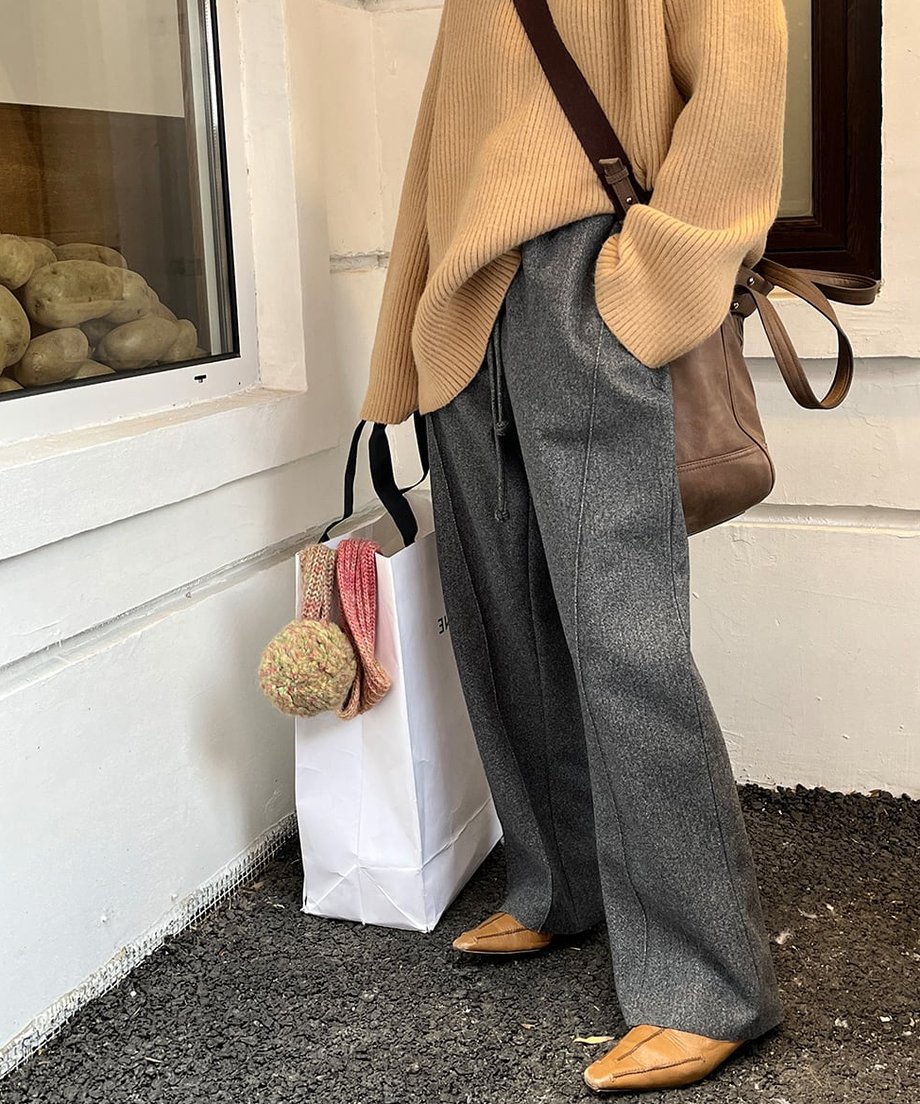 ダークグレーのセンターシームワイドパンツを着用した女性の全身。キャメル色のニットと革製バッグ、ポインテッドトゥのフラットシューズを合わせたスタイル。