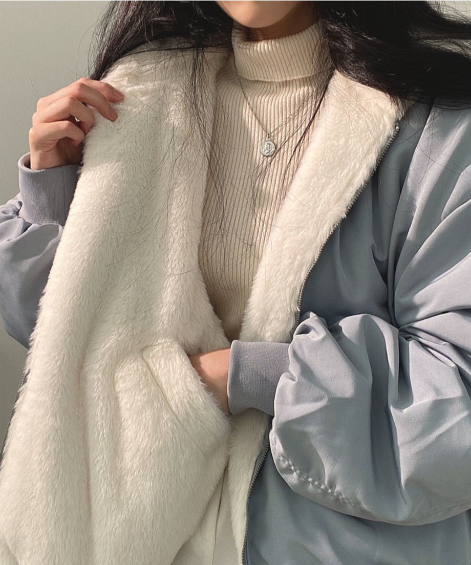 ブルーのブルゾンを着用した女性が、内側の白いボアを広げて見せている上半身のクローズアップ。白いタートルネックニットにネックレスを着用。