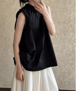 黒のノースリーブチュニックブラウスと、白のロングスカートを合わせた女性のコーディネート。ブラウスの首元はフリルデザインで、女性がスマホを持って顔の一部を隠している。