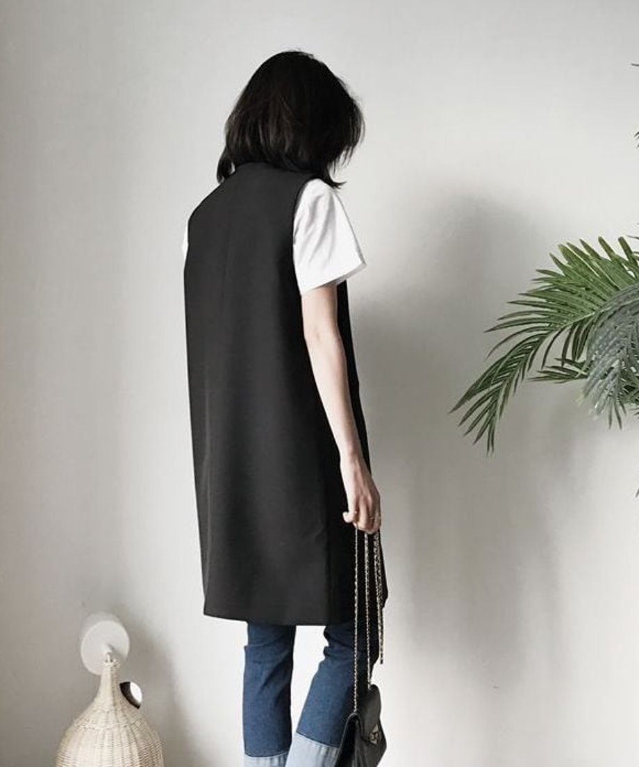 後ろ姿の女性が、白い半袖Tシャツとデニムの上から黒のロング丈テーラードベストを着用し、黒のチェーンバッグを持っている。