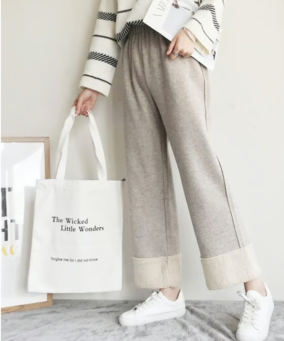 明るいグレージュのワイドパンツを着用した女性の全身画像。パンツの裾はオフホワイトのボア（フリース）素材を折り返しており、白いボーダー柄のセーターを着用し、白いスニーカーと「The Wicked Little Wonders」と書かれた白いトートバッグを持っている。