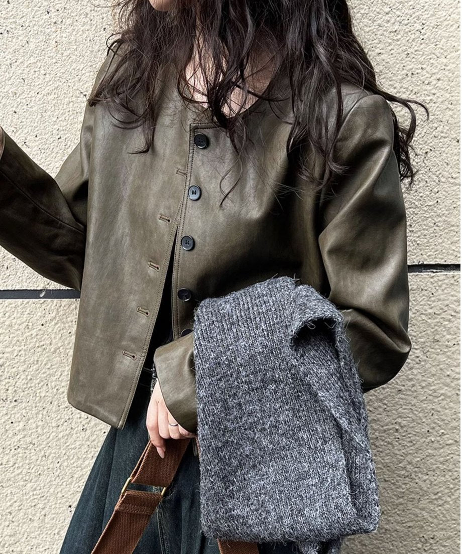 カーキのフェイクレザージャケットを着用した女性のバストアップ。左腕にグレーのニットをかけ、茶色のバッグを持っている。
