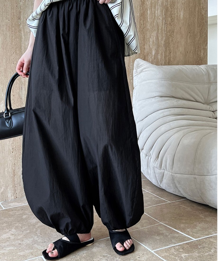 ブラックのバルーンパンツ（裾絞り）とストライプ柄のトップスを着用し、黒いバッグを持った女性の、屋内の全身