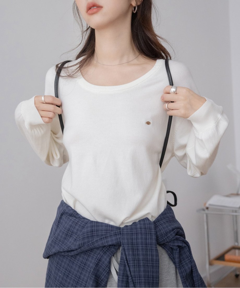 ホワイトのUネックニットを着用した女性のバストアップ。細い黒いストラップを肩にかけ、ネイビーのチェックシャツを腰に巻いている。