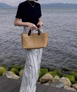 ホワイトストライプ柄のボトムスを履いた女性が黒いトップスと籠バッグを持ち、海辺を歩いている様子。