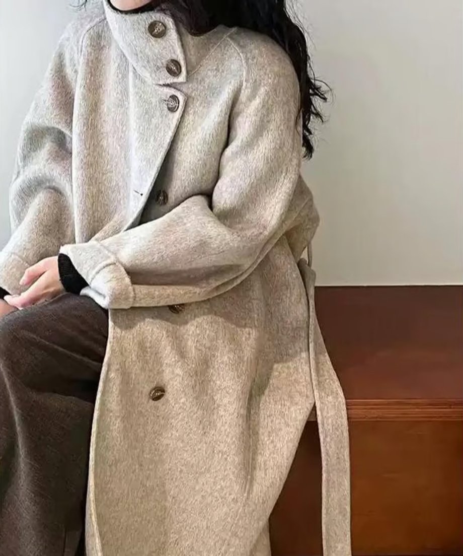 モカのスタンドカラーコートを着用した女性が茶色のベンチに座っている上半身。襟元のボタンとワイドな袖口が見える。